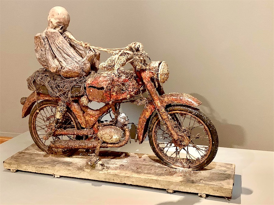 composition réalisée avec une vieille moto rouillée, une momie en position foetale sur le siège.