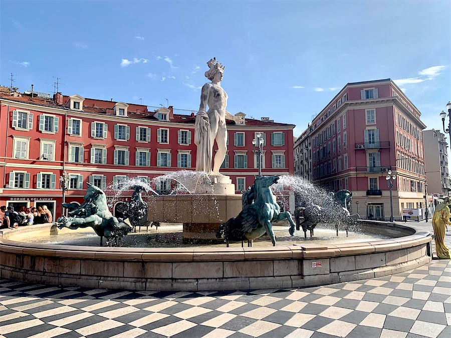 place ronde, bordée d'immeubles suivant l'arrondi, façades rouges, fenêtres à persiennes claires. Au premier plan, fontaine ronde, statue de marbre au centre et chevaux de bronze autour.