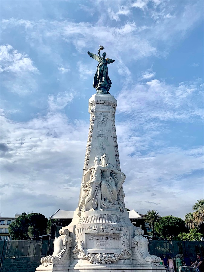 statue représentant une Victoire ailée en bronze, posée sur une base en marbre en forme d'obélisque. Ciel bleu parsemé de légers nuages.