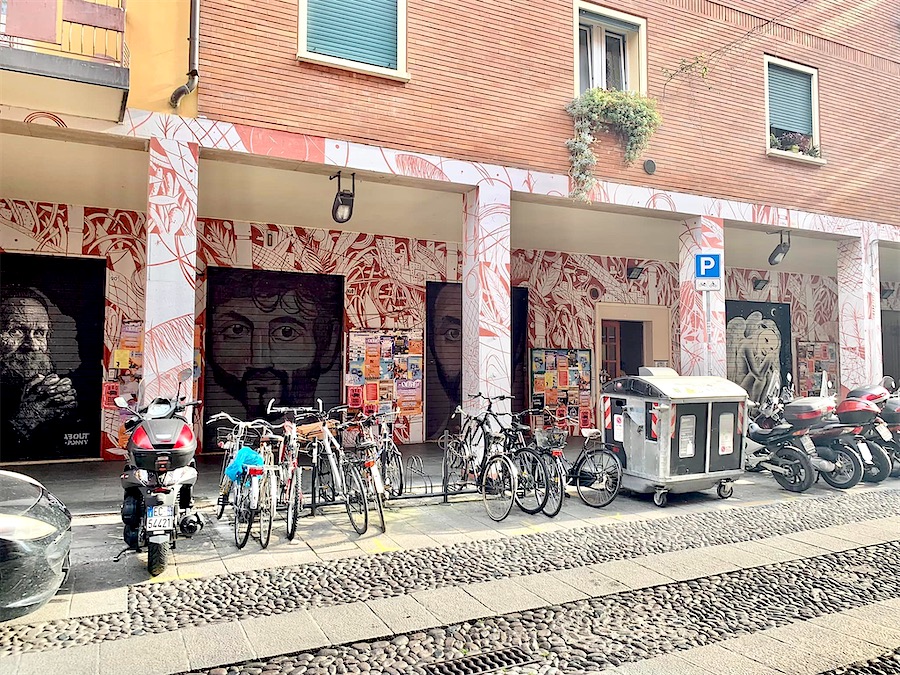 rue de Bologne, nombreux deux roues stationnés. À l'arrière-plan, mur de façade du rez de chaussée du batiment et portiques décorés en blanc et rouge et portraits en noir et blanc