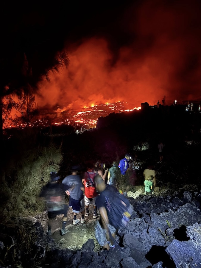 au premier plan, groupe de personnes marchant sur la roche et au second plan, lave en fusion et ciel rougeoyant sous les nuages de vapeur